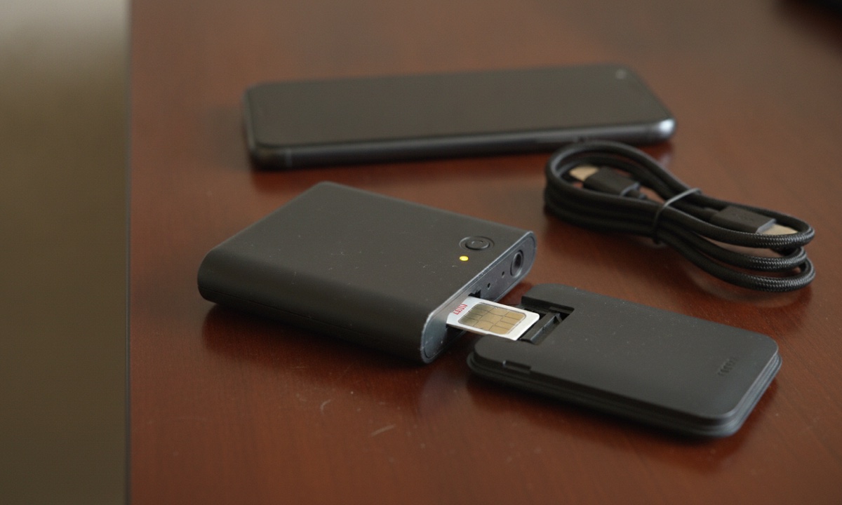 A pocket Wi-Fi router with its back cover open and SIM tray exposed on a desk, phone resting nearby
